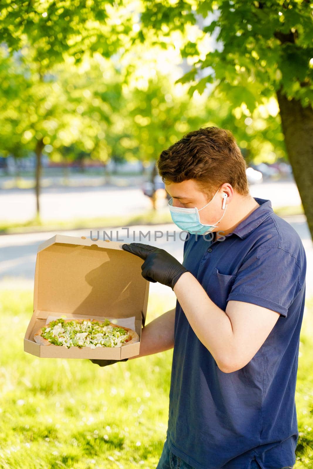 Courier in medical mask and gloves holds pizza in hands and looks to pizza with green salad, tomatoes, cheese