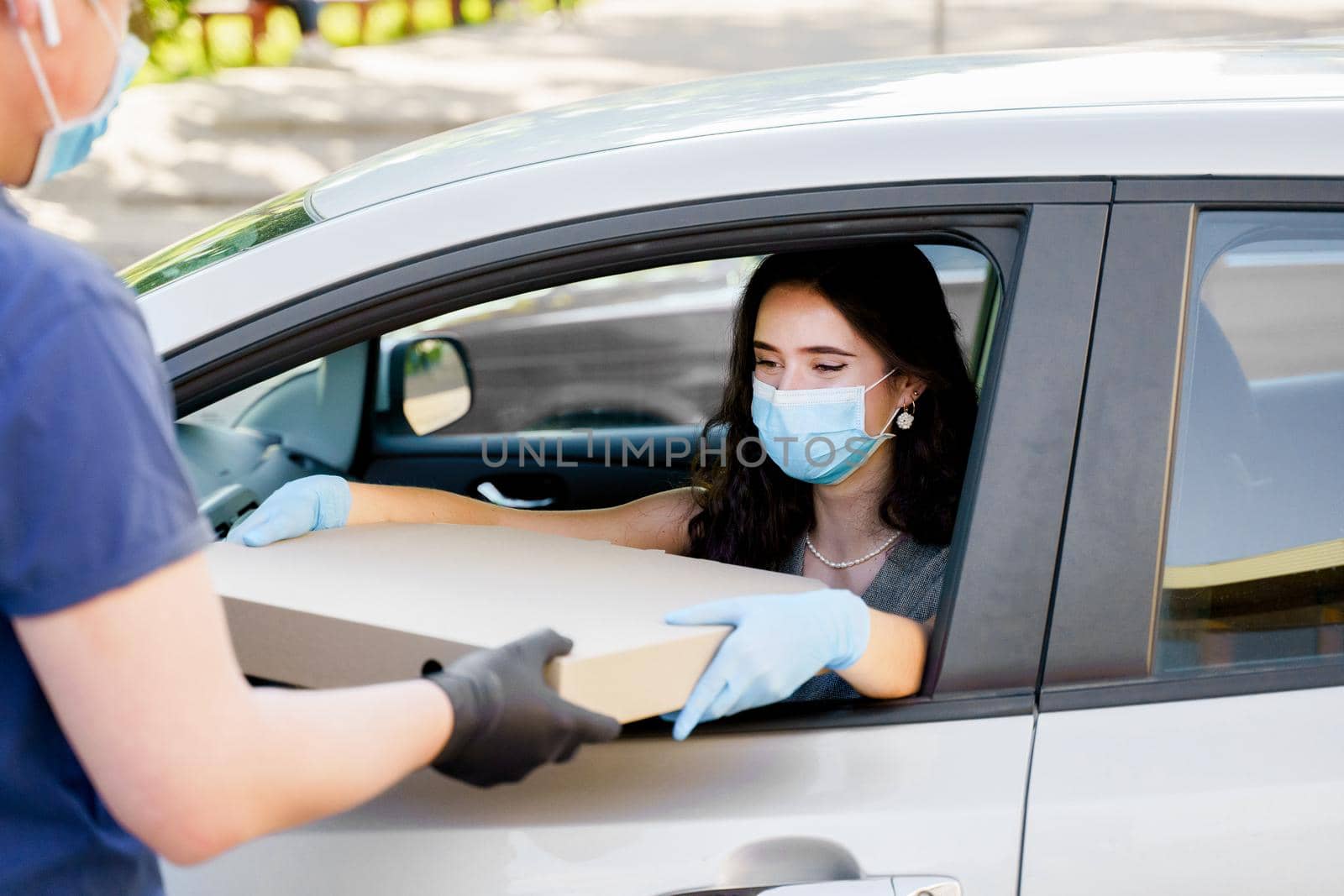 Girl in medical mask and gloves gets huge pizza in car. Big size pizza in carton eco box. Courier gives large pizza to young business woman. Delivery by car. by Rabizo