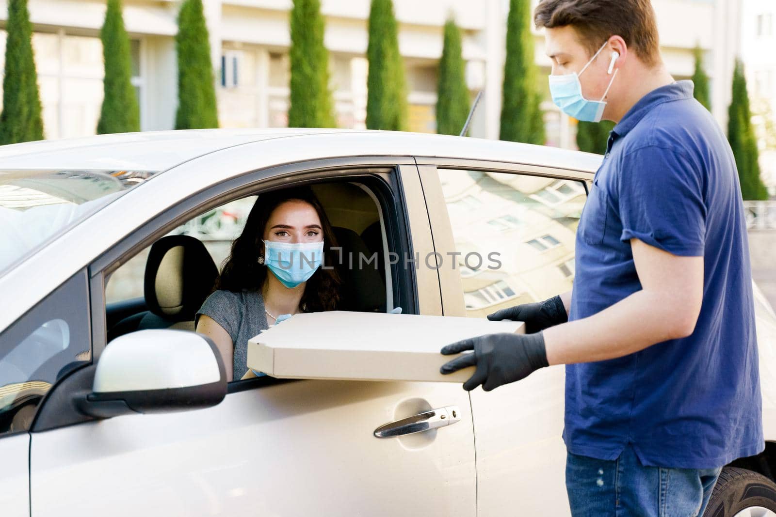 Girl in medical mask and gloves gets huge pizza in car. Big size pizza in carton eco box. Courier gives large pizza to young business woman. Delivery by car. by Rabizo