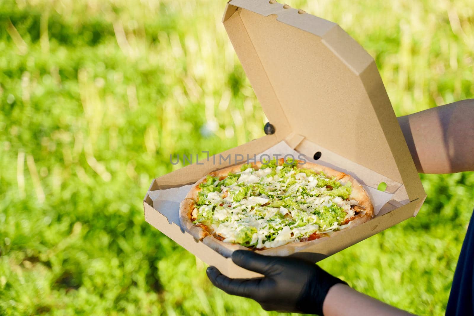 Pizza close-up in hands in medical gloves. Courier in medical mask holds pizza in hands and looks to pizza with green salad, tomatoes, cheese