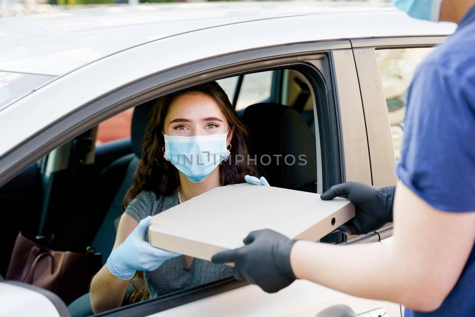 Food delivery courier gives pizza to woman in eco car. Tasty pizza with salami, cheese, tomatoes in cardoard box. Giving food trought car window. by Rabizo