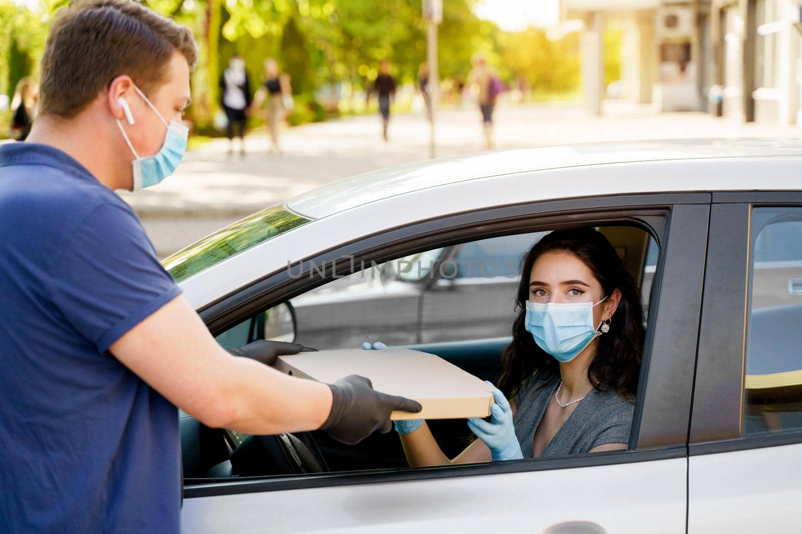 Safe food delivery from pizzeria to car during quarantine coronavirus. Attractive business woman in medical mask and gloves gets pizza in cardboard box. by Rabizo