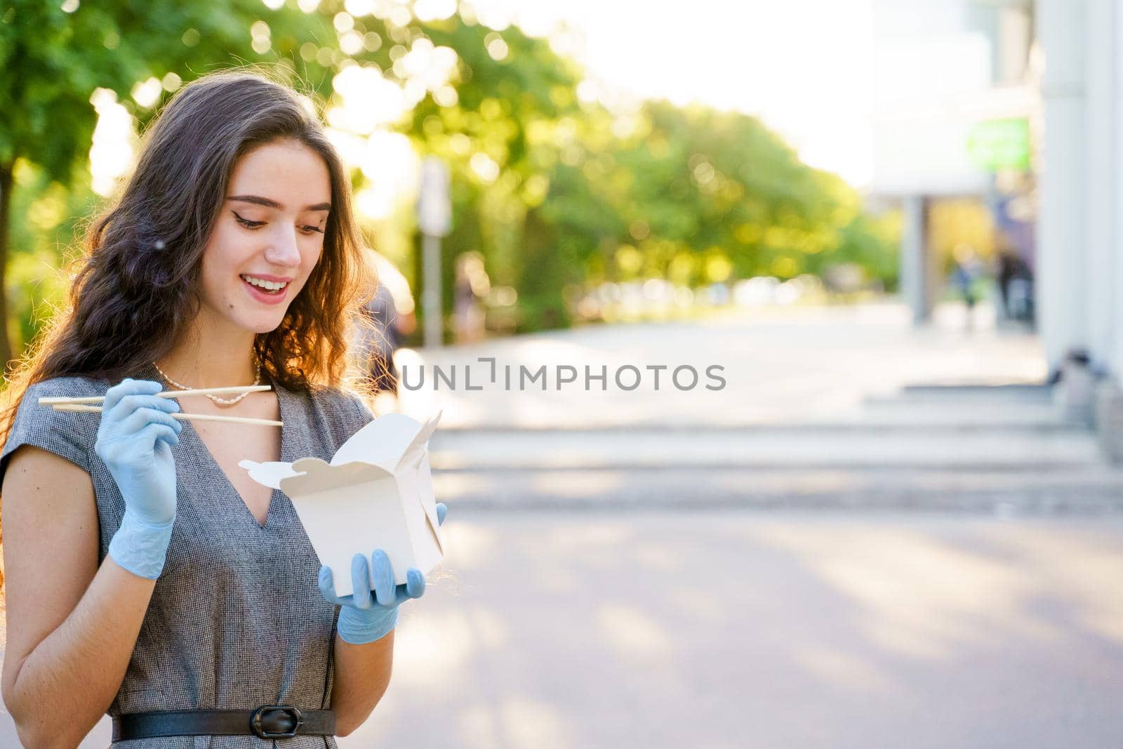 Young surprised girl with curly hair in medical gloves and mask holds wok in box udon noodles in hands and smiles. Udon noodles in white box delivery by Rabizo