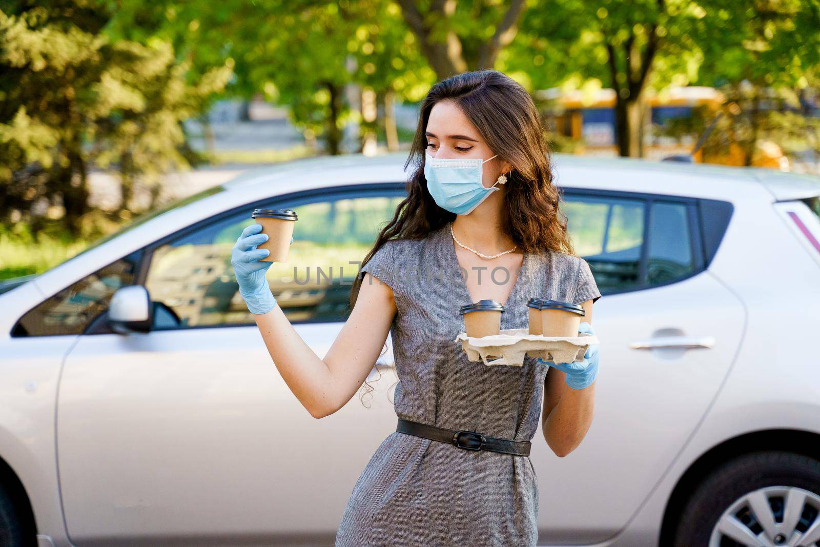 Girl in medical mask holds 4 coffee in hands with gloves. One cup of coffee in hands. Delivery by car. Safe delivery by Rabizo