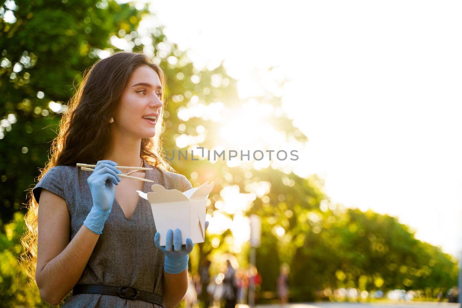 Young surprised girl with curly hair in medical gloves and mask holds wok in box udon noodles in hands and smiles. Udon noodles in white box delivery.