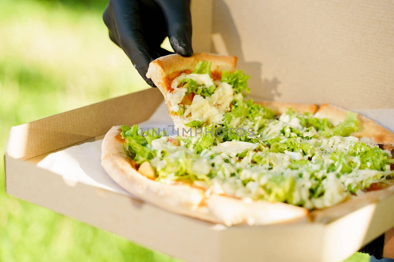 Pizza close-up in hands in medical gloves. Courier in medical mask holds pizza in hands and looks to pizza with green salad, tomatoes, cheese