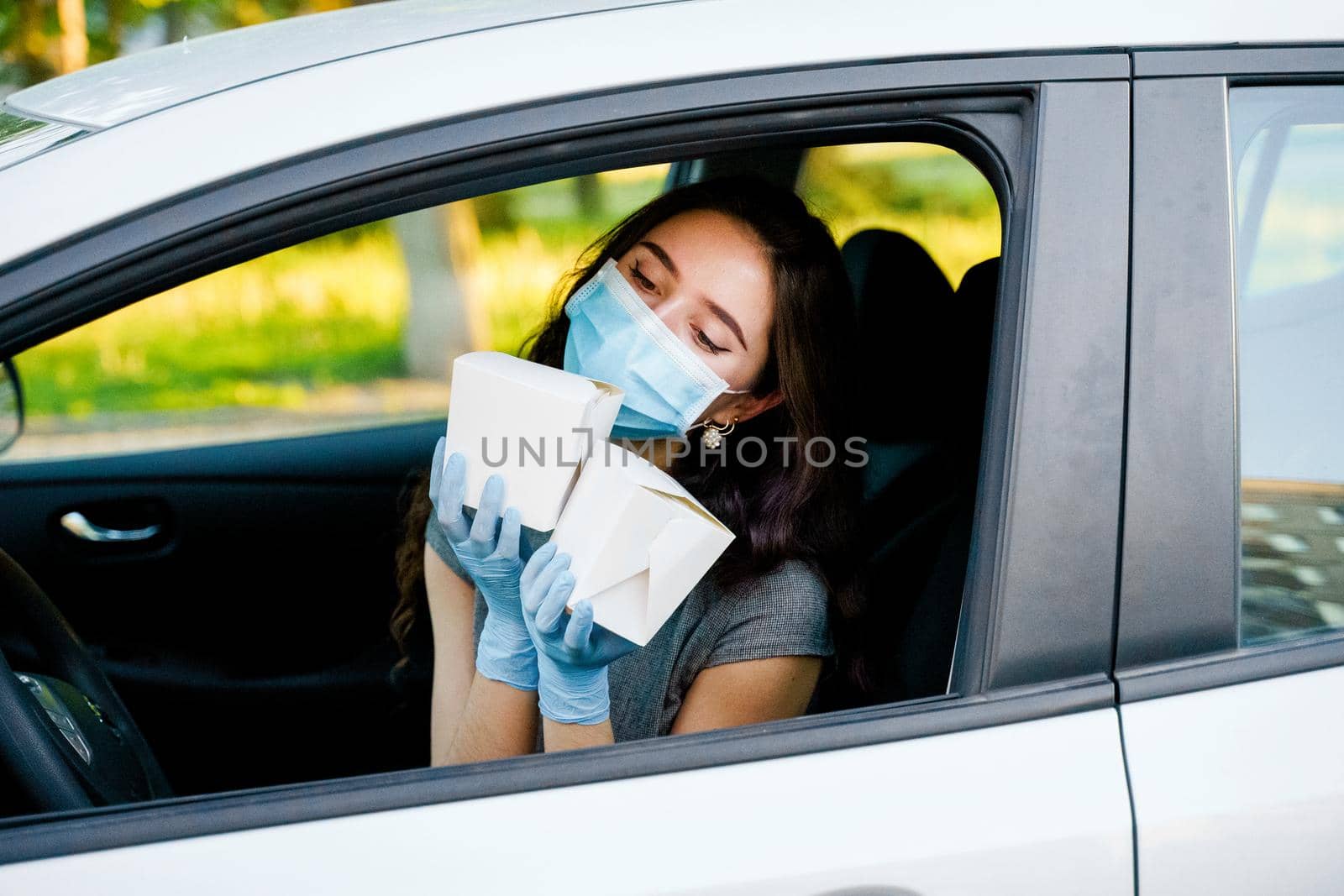 Young attractive girl in car in medical gloves and mask holds wok in box udon noodles in hands and smiles. Udon noodles in white box delivery