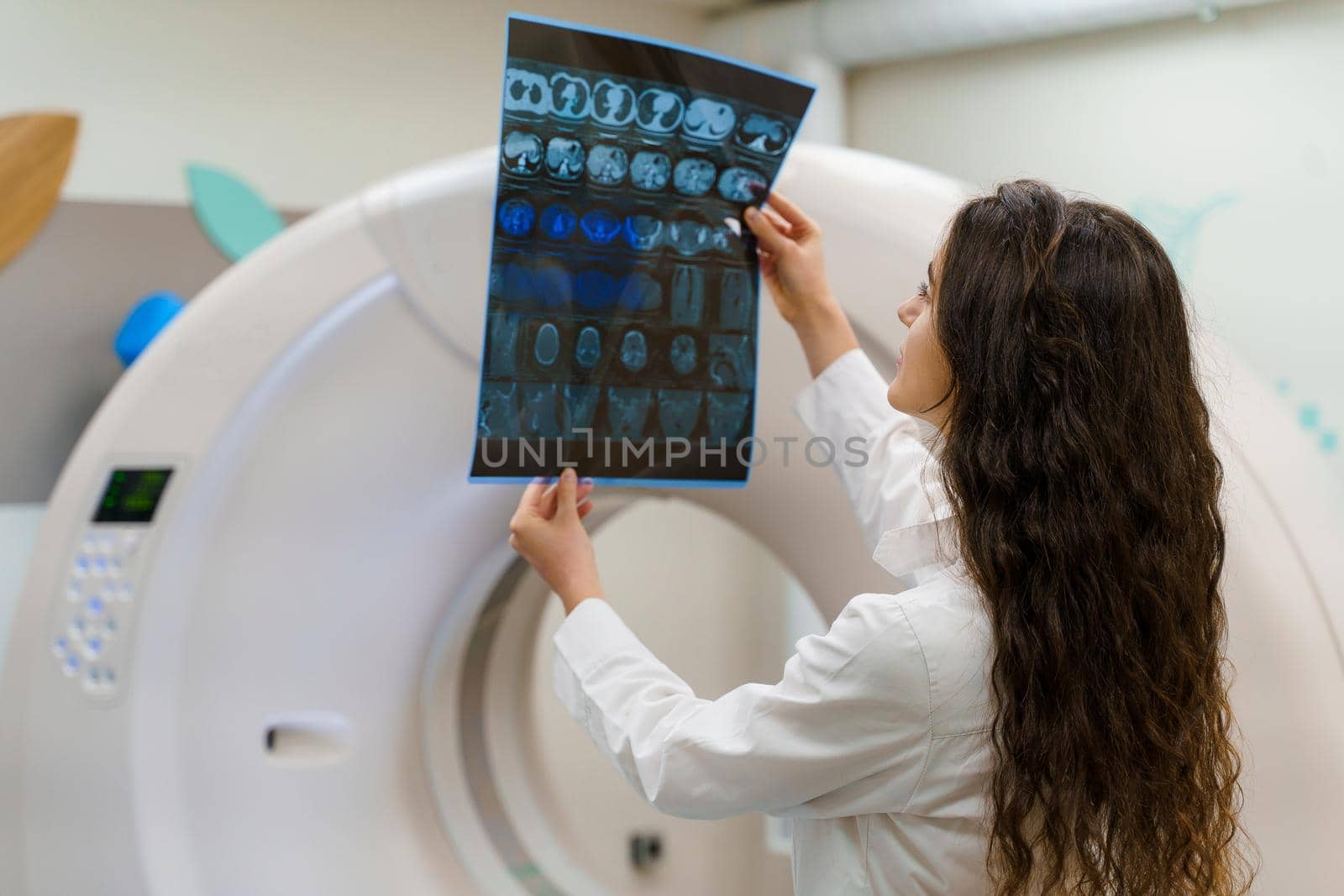 Young caucasian girl doctor looks at x-ray picture of patient brain near kt komputer tomograhphy. Young girl in medical clinic gotes new expirience. by Rabizo