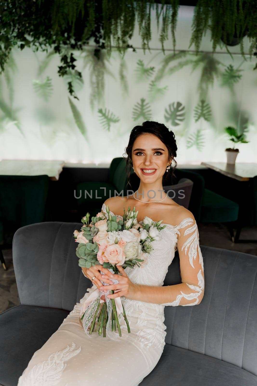 Bride with wedding bouquet smiles, looks in camera and touches her face. Attractive girl portrait. Girl in wedding dress in luxury restaurant