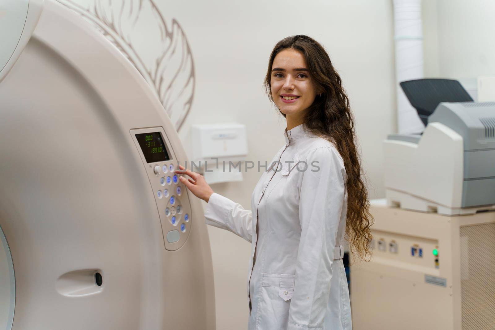Medical doctor does computer tomography fot patient. Young girl in white coat looks in camera and press button on CT