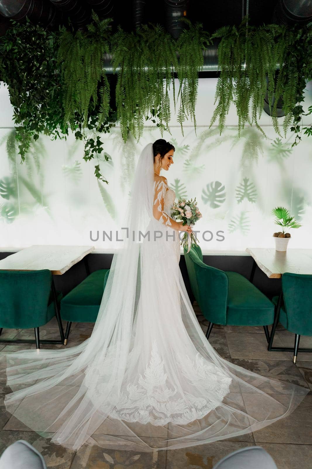 Young attractive bride in wedding dress with bridal veil looks at bouquet and smiles. Girl in wedding gown in modern restaurant. by Rabizo