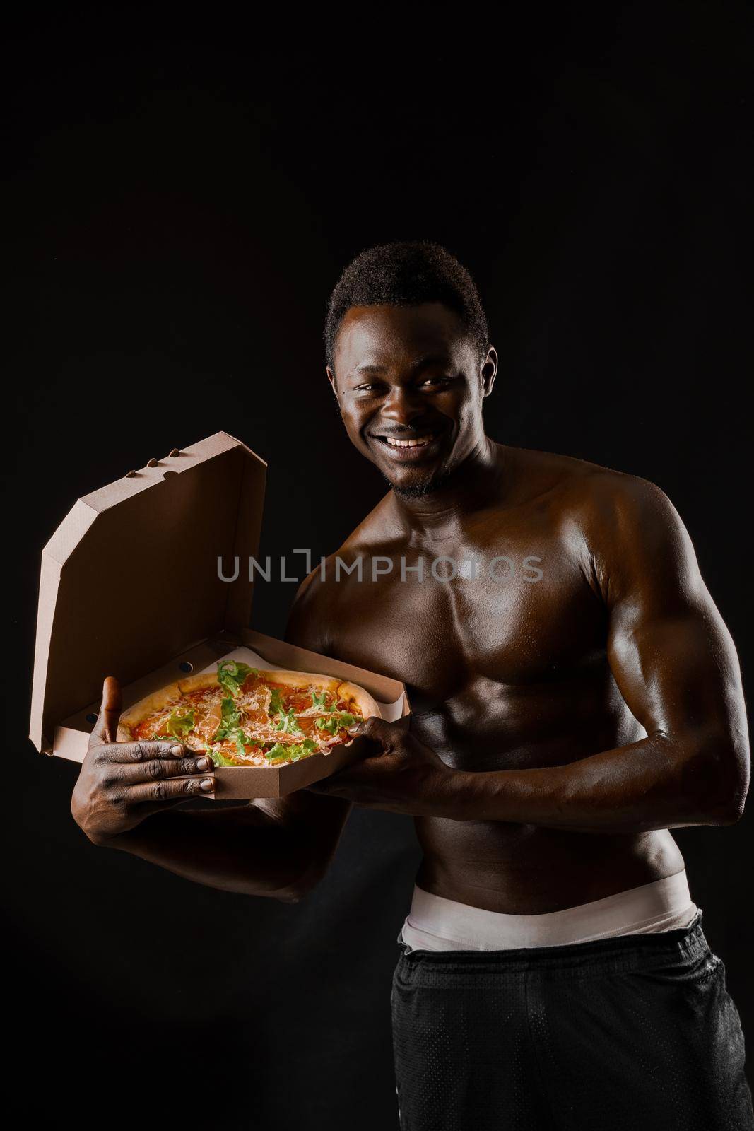Athletic handsome black man works in pizza delivery. Stay at home and eat a pizza. Tasty food. African man smiles and shows a pizza in cardboard box by Rabizo