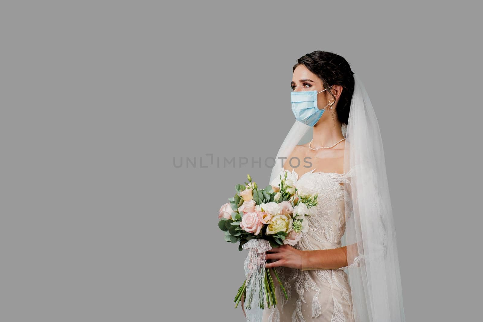 Bride in medical mask and wedding bouquet at coronavirus covid-19 quarantine period. Attractive girl in studio.