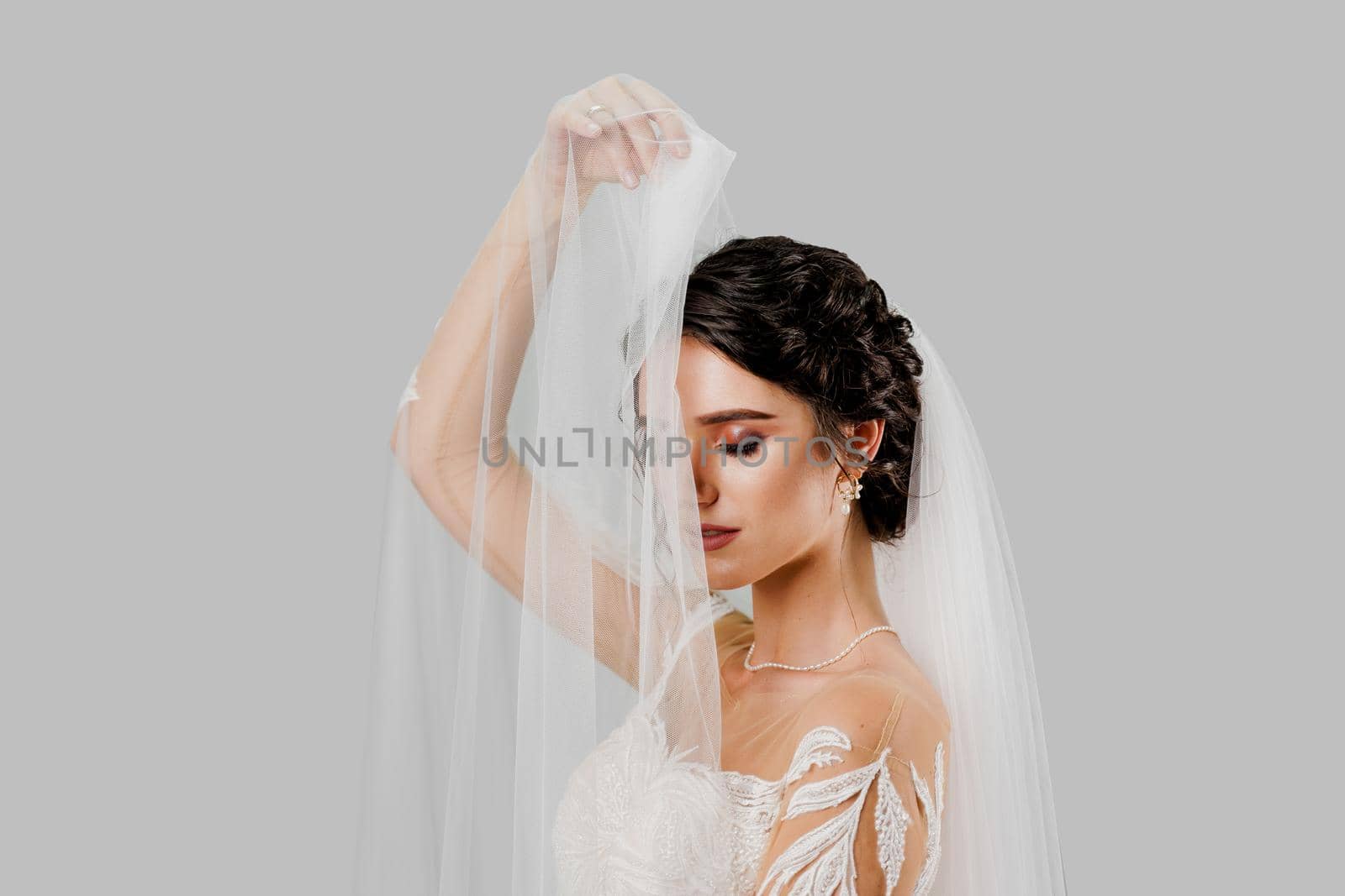 Young attractive bride in wedding dress touches bridal veil on white background in studio. Advert for social networks for wedding agency and bridal salon.