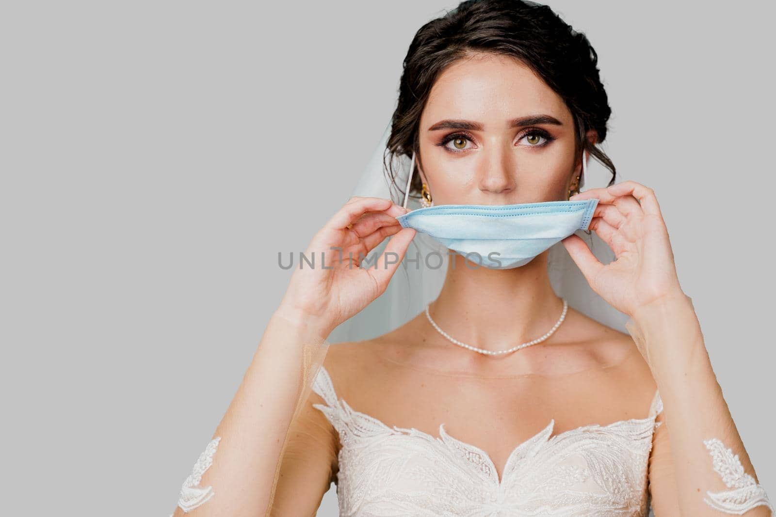 Bride in medical mask and wedding bouquet at coronavirus covid-19 quarantine period. Attractive girl takes off mask and looks in camera in studio