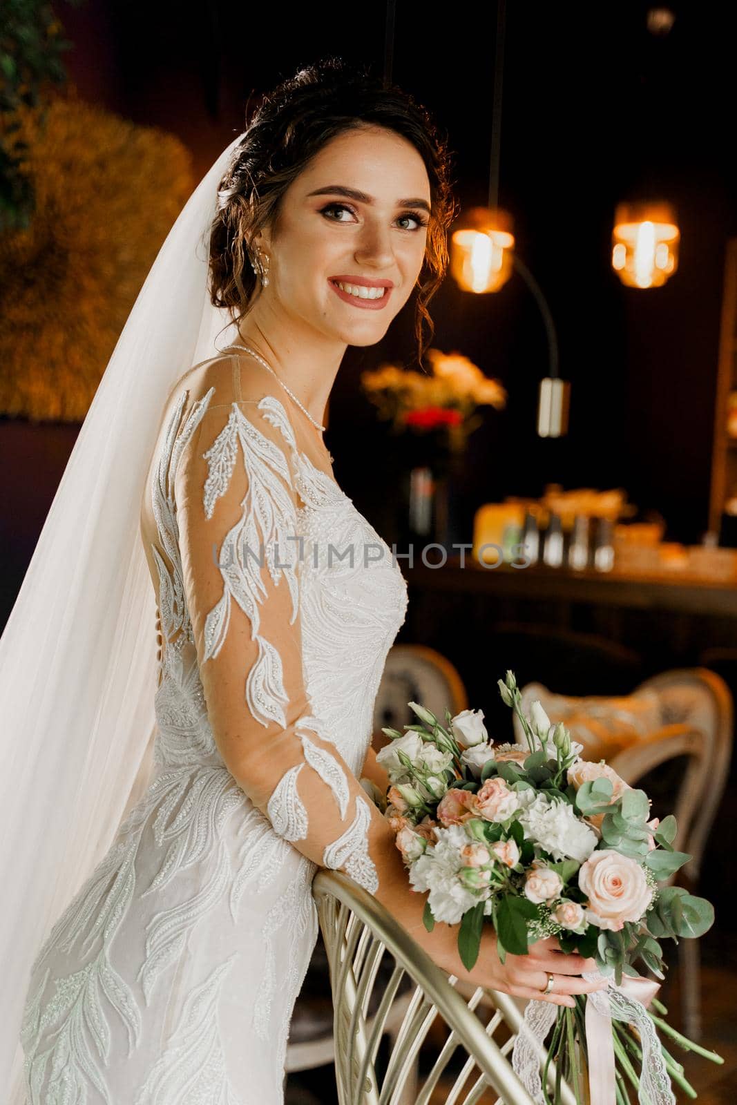 Bride with wedding bouquet smiles, looks in camera and touches her face. Attractive girl portrait for social networks. Girl in wedding dress in luxury restaurant. by Rabizo