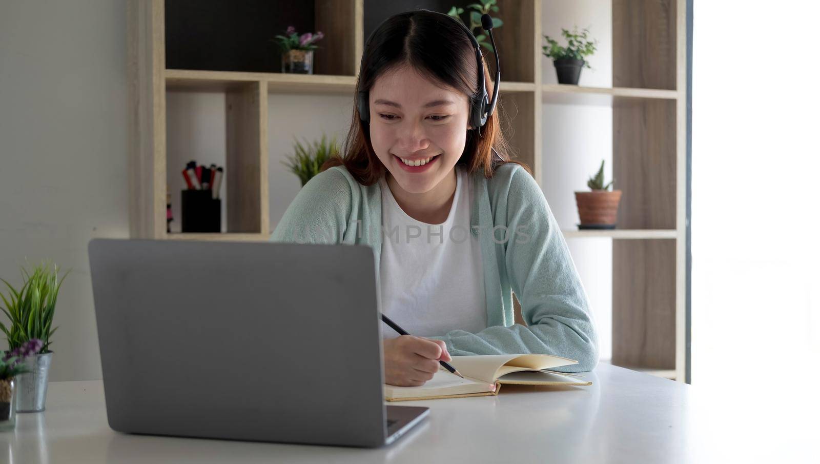 Happy young woman in headphones speaking looking at laptop making notes, girl student talking by video conference call, female teacher trainer tutoring by webcam, online training, e-coaching concept by wichayada