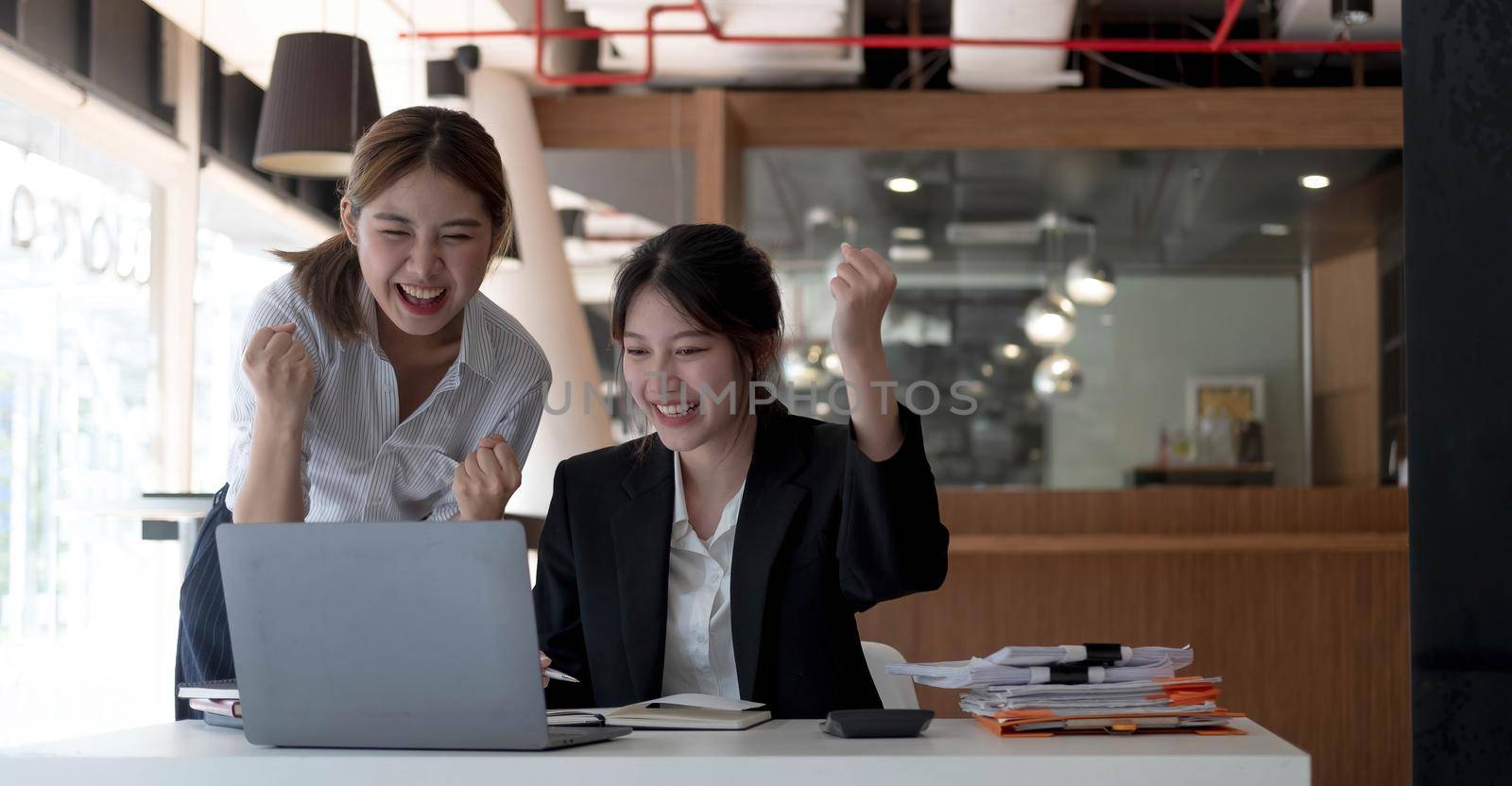 Two young Asian businesswomen show joyful expression of success at work smiling happily with a laptop computer in a modern office..