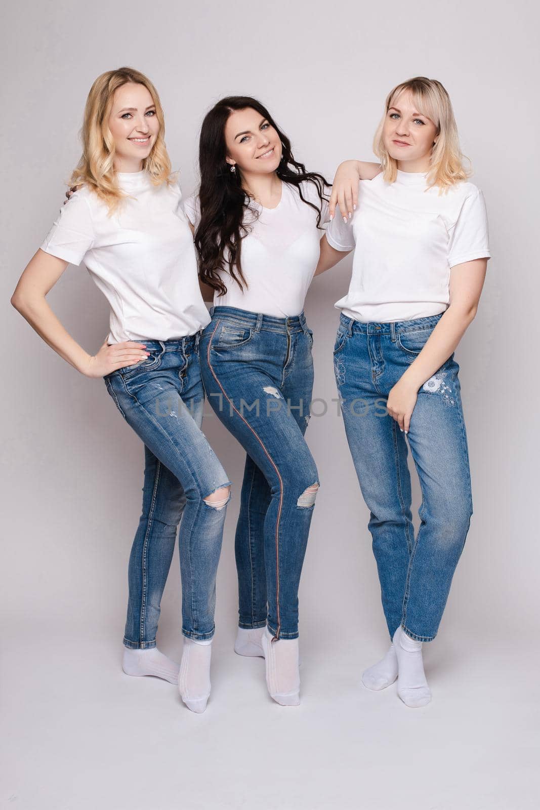 Front view of three beautiful women wearing white shirts and jeans and posing on isolated background. Stylish models standing in studio, looking at camera and smiling. Concept of casual street outfit.