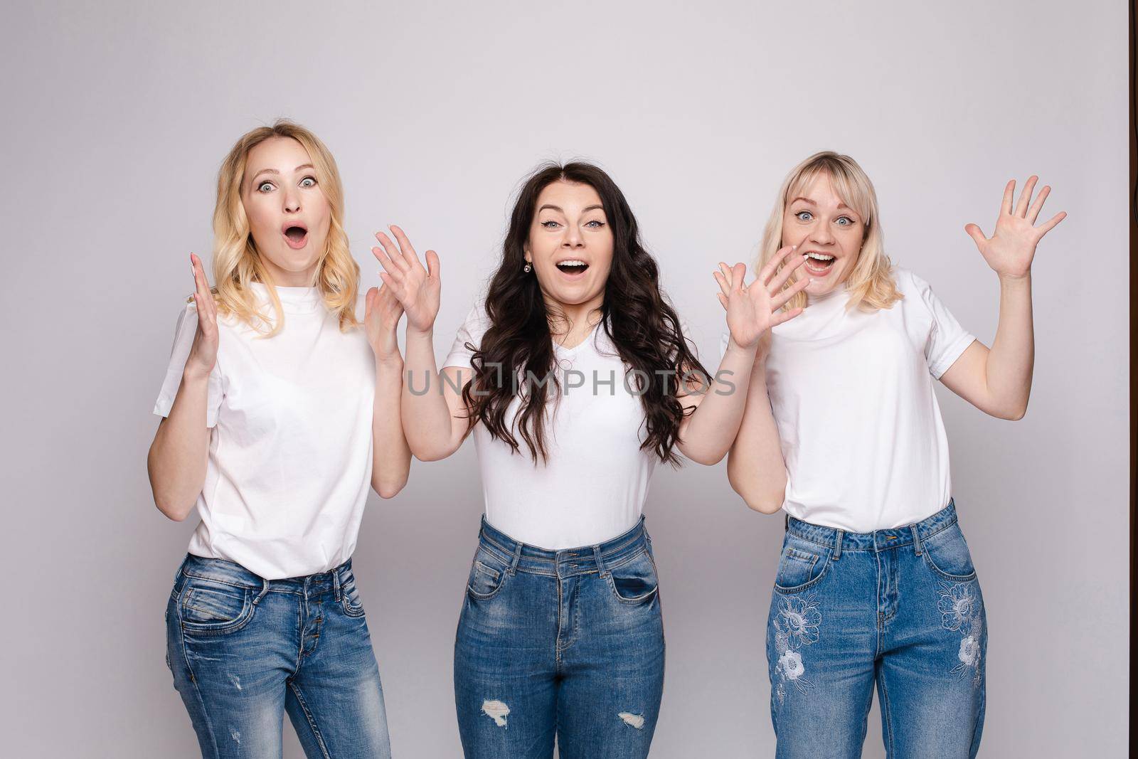 Three female friends looking at camera and shouting in surprise on white isolated background in studio. Amazed girl in white shirts and jeans with big eyes and open mouth posing. Concept of emotions.