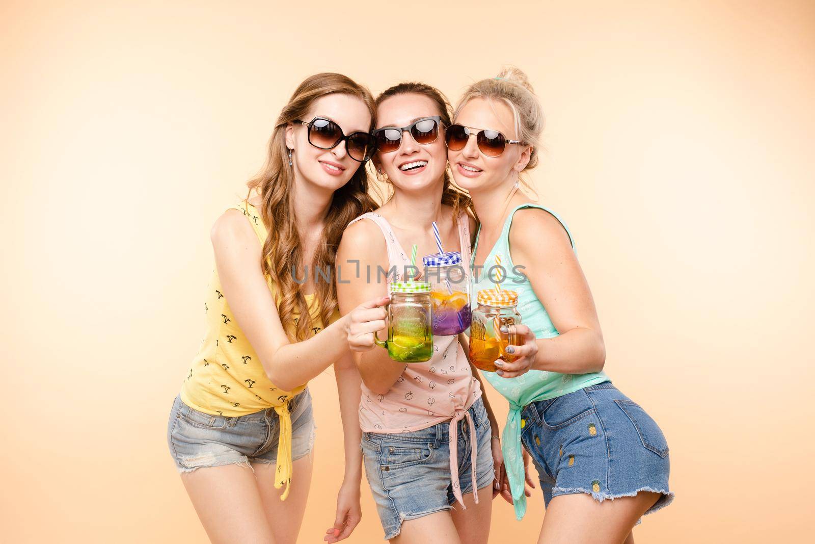 Happy young girls standing close to each other and holding chilling drinks. Group of friends in casual clothes drinking lemonades during holidays. Beautiful woman in sunglasses posing with cocktails.