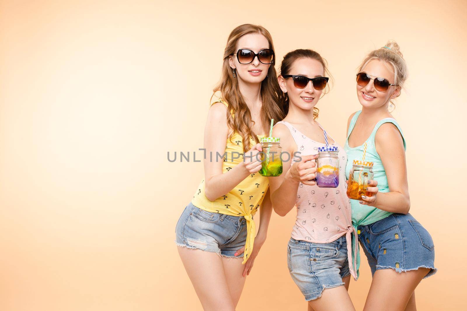 Three sporty girls in tops, shorts and glasses keeping fresh juice and posing on isolated background. Happy young women laughing and caring about body and fit. Concept of energy and shape.