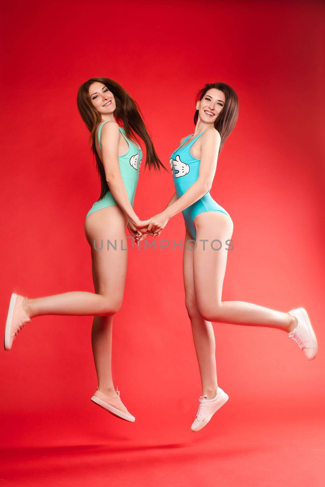 Side view of young models in swimming wear jumping together and laughing on red isolated background. Two active girl with long legs looking at camera and posing. Concept of fit and body.