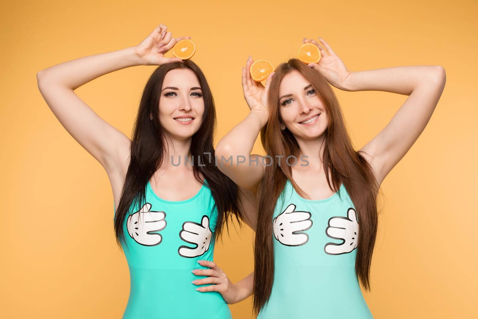 Front view of female friends keeping fresh oranges and posing on isolated background in studio. Beautiful young girls looking at camera and laughing while keeping fruits. Concept of health.