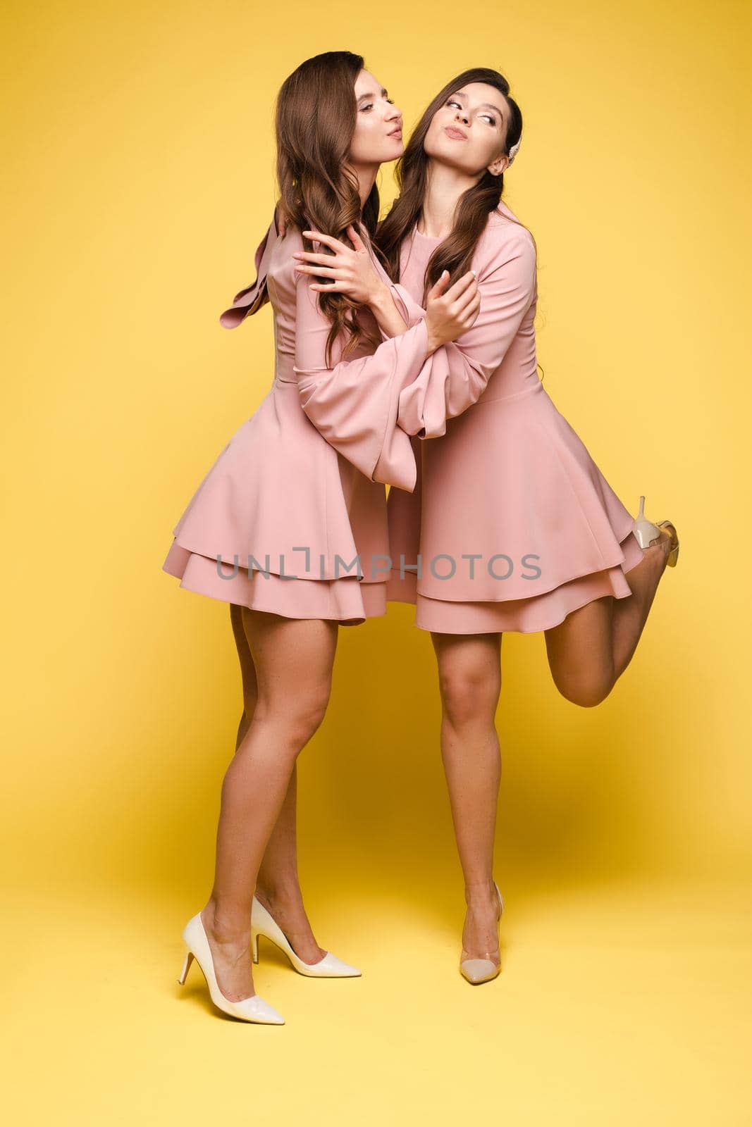 Brunette twins with long hair in pink dresses posing at camera on yellow studio background. Happy sisters in elegant clothes smiling and standing together. Beautiful girl leaning on her friend.