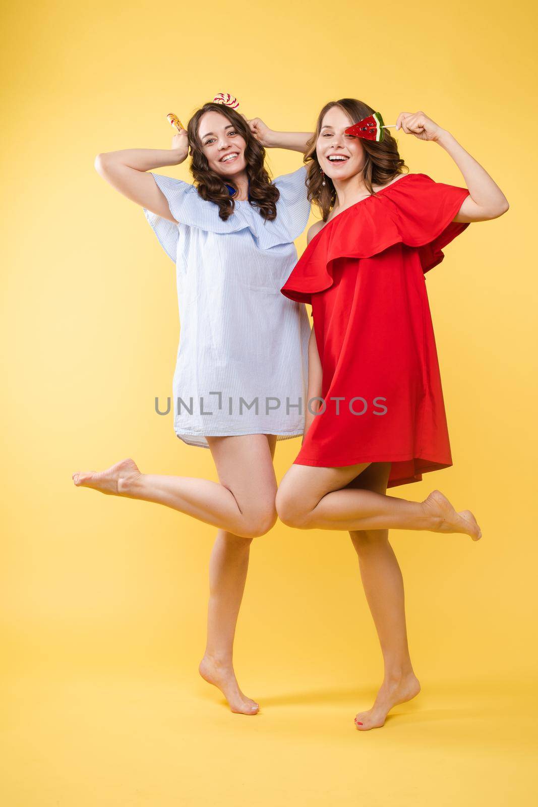 Cute brunette sisters in red and blue dresses over yellow background. by StudioLucky