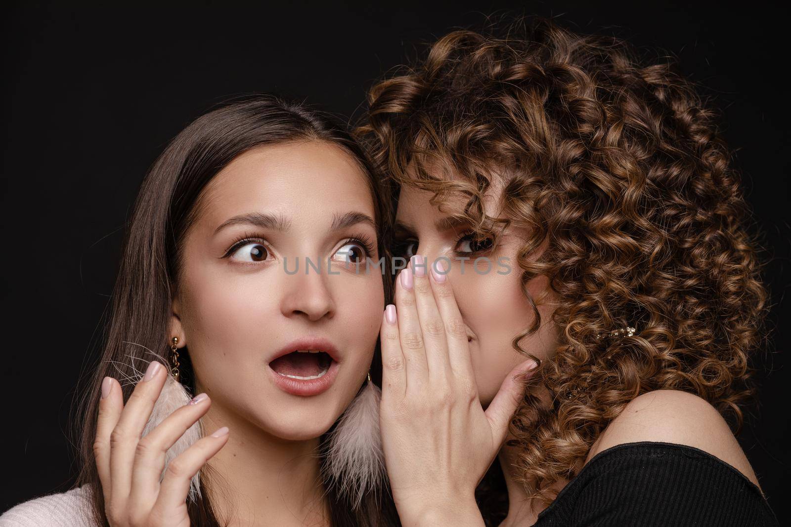 Front view of shocked brunette with big eyes and open mouth in white dress looking at camera while female friend whispering secret on black isolated background. Young women gossiping in studio.