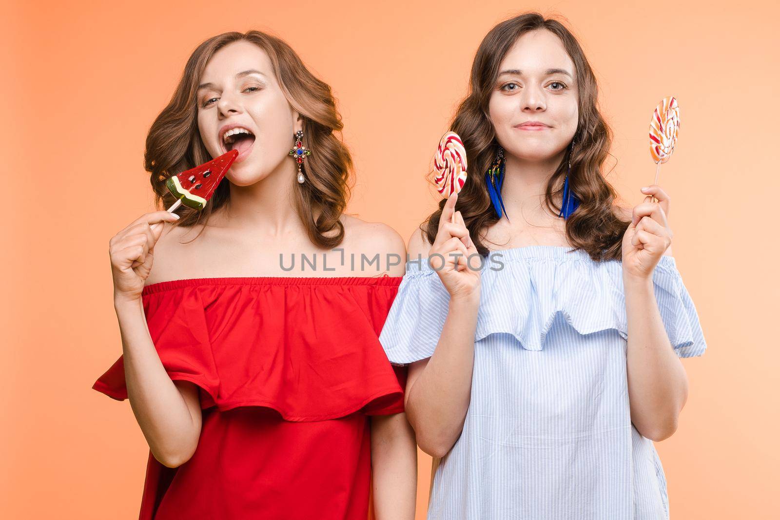 Front view of charming twins in pink dresses closing eyes with lollipops and posing on isolated background. Two female friends laughing and having fun in studio. Concept of childhood and beauty.