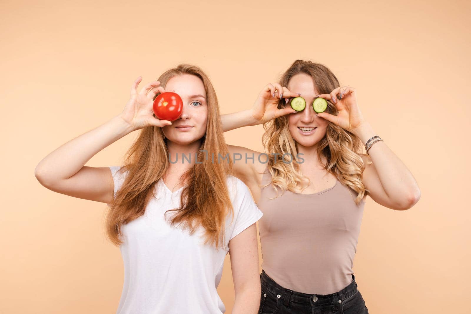 Front view of two female friends keeping fresh healthy tomatoes and cucumbers on isolated background. Happy girls keeping vegetables and fruits, posing and laughing in studio. Concept of diet.