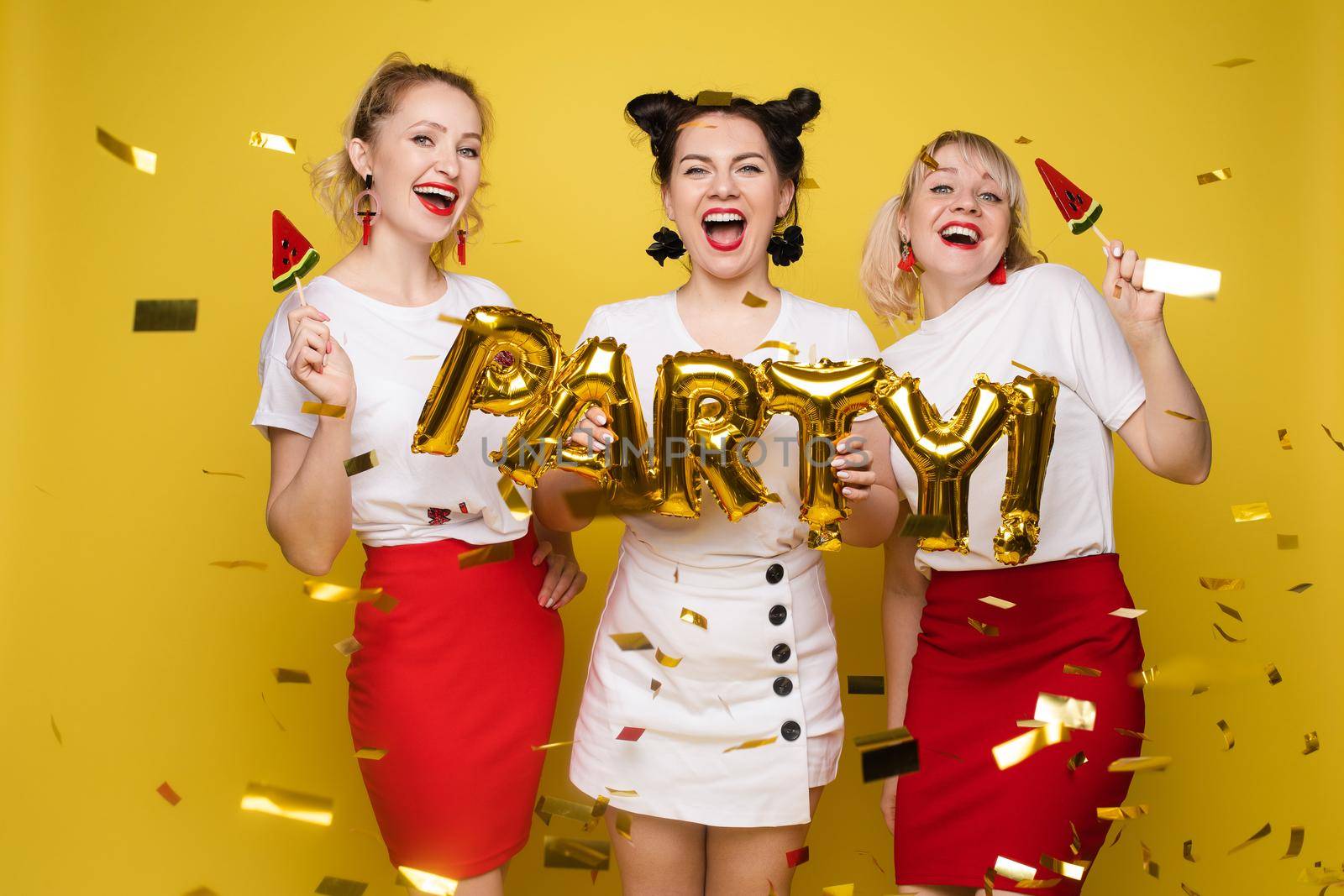 Front view of bright three girls in white shirts keeping balloons and ice creams on yellow isolated background. Cheerful pretty women looking at camera and posing. Concept of party and sparkles.