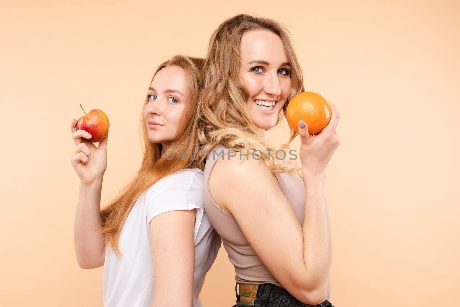 Side view of two beautiful friends standing back to back and looking at camera with smile. Girl on the right holding bright juicy orange. Healthy food concept.
