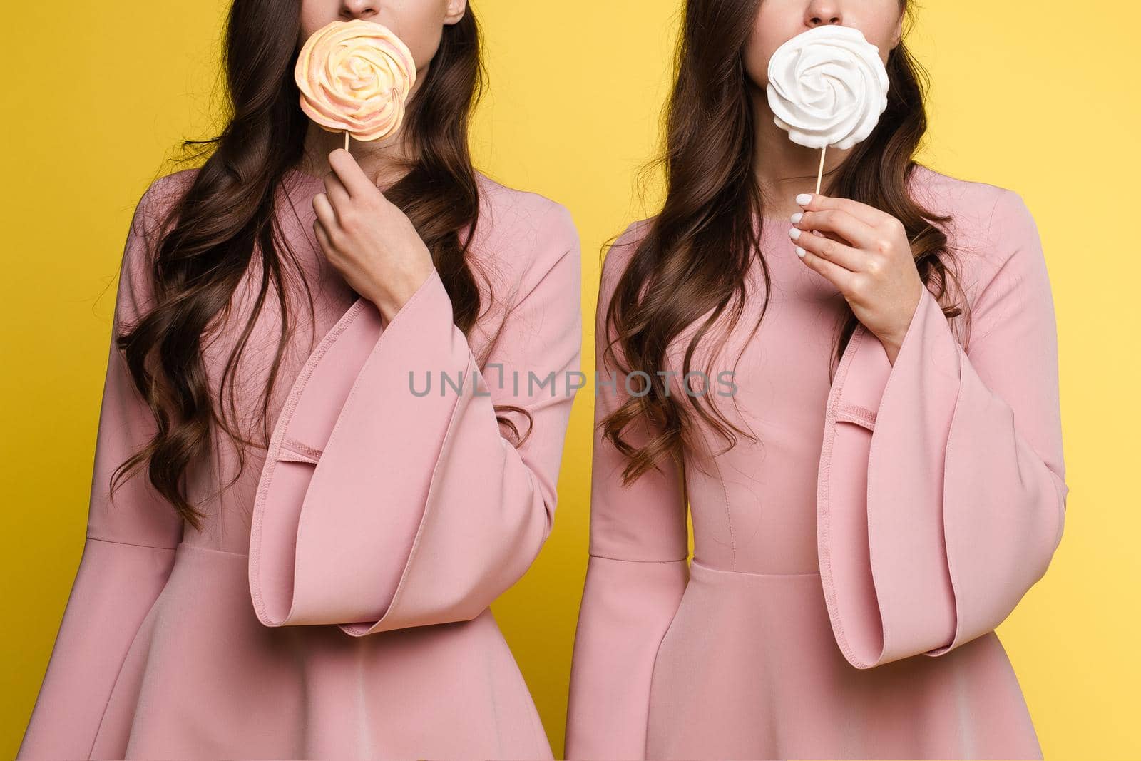 Front view of charming twins in pink dresses closing eyes with lollipops and posing on isolated background. Two female friends laughing and having fun in studio. Concept of childhood and beauty.