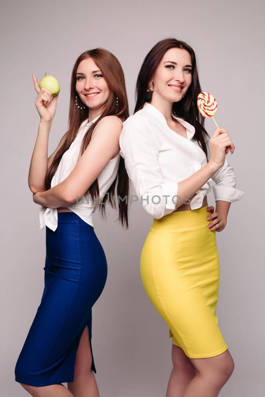 Studio portrait of two gorgeous young brunette sisters or friends or colleagues in blue and yellow skirts and white blouses posing smiling at camera. One is holding a healthy apple and other is sweet candy.