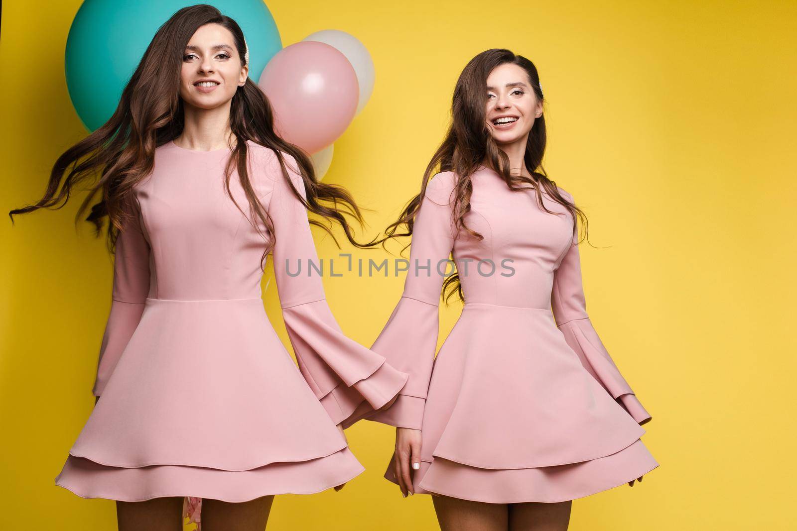 Studio portrait of gorgeous brunette twins posing in trendy elegant pink dresses. Stylish outlooks. Smiling at camera. Yellow background with mint and pink air balloons.