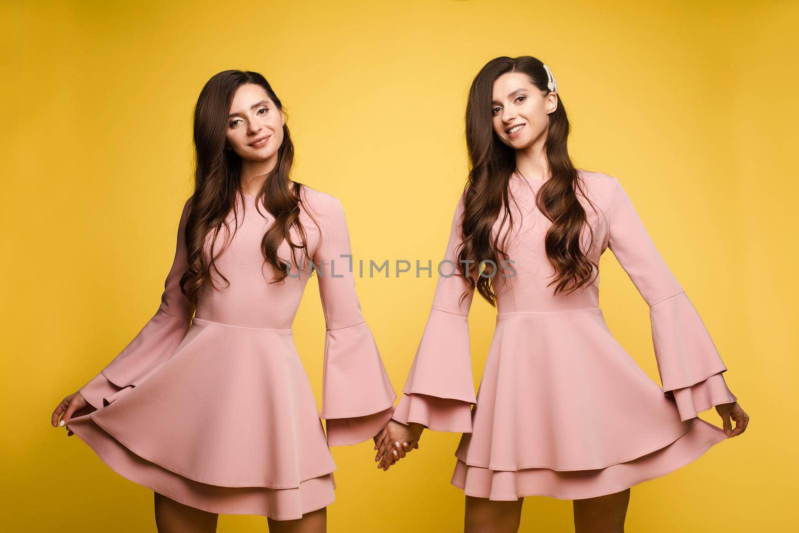 Brunette twins with long hair in pink dresses posing at camera on yellow studio background. Happy sisters in elegant clothes smiling and standing together. Beautiful girl leaning on her friend.