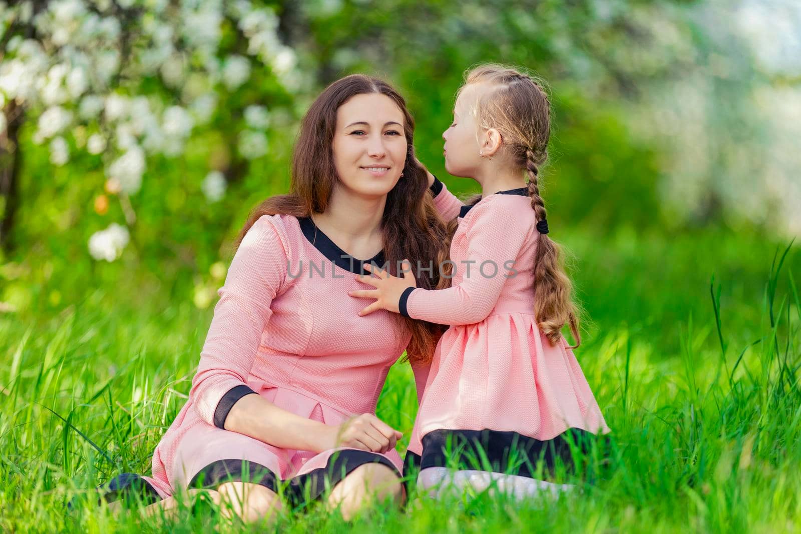 mother and daughter sit in the green grass against the backdrop of blooming apple trees