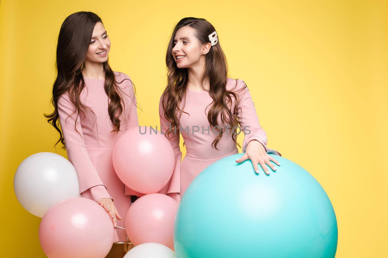 Cute brunette twins in pink dresses over yellow background. by StudioLucky