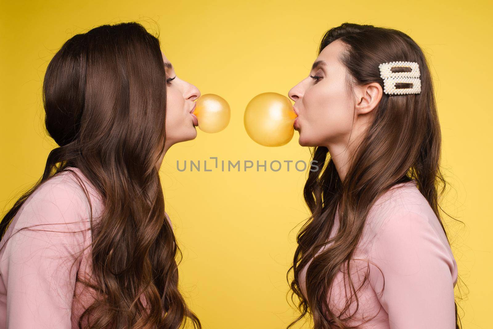 Young twins in elegant dresses holding pink and white balloons in their hands. Brunette sisters with long hair blowing big bubbles from chewing gum. Beautiful girls posing at camera with decorations.