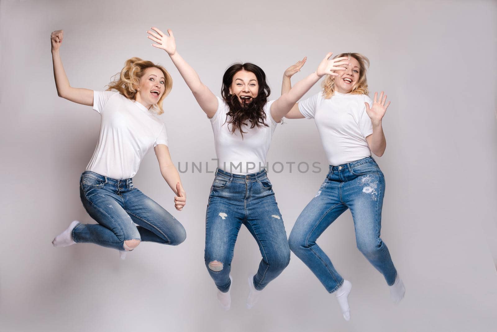 Close up of two blonde and one brunette women getting fun together. Three friends in casual clothes dancing and jumping. Beautiful happy girls in white t-shirts and jeans smiling and hanging out.