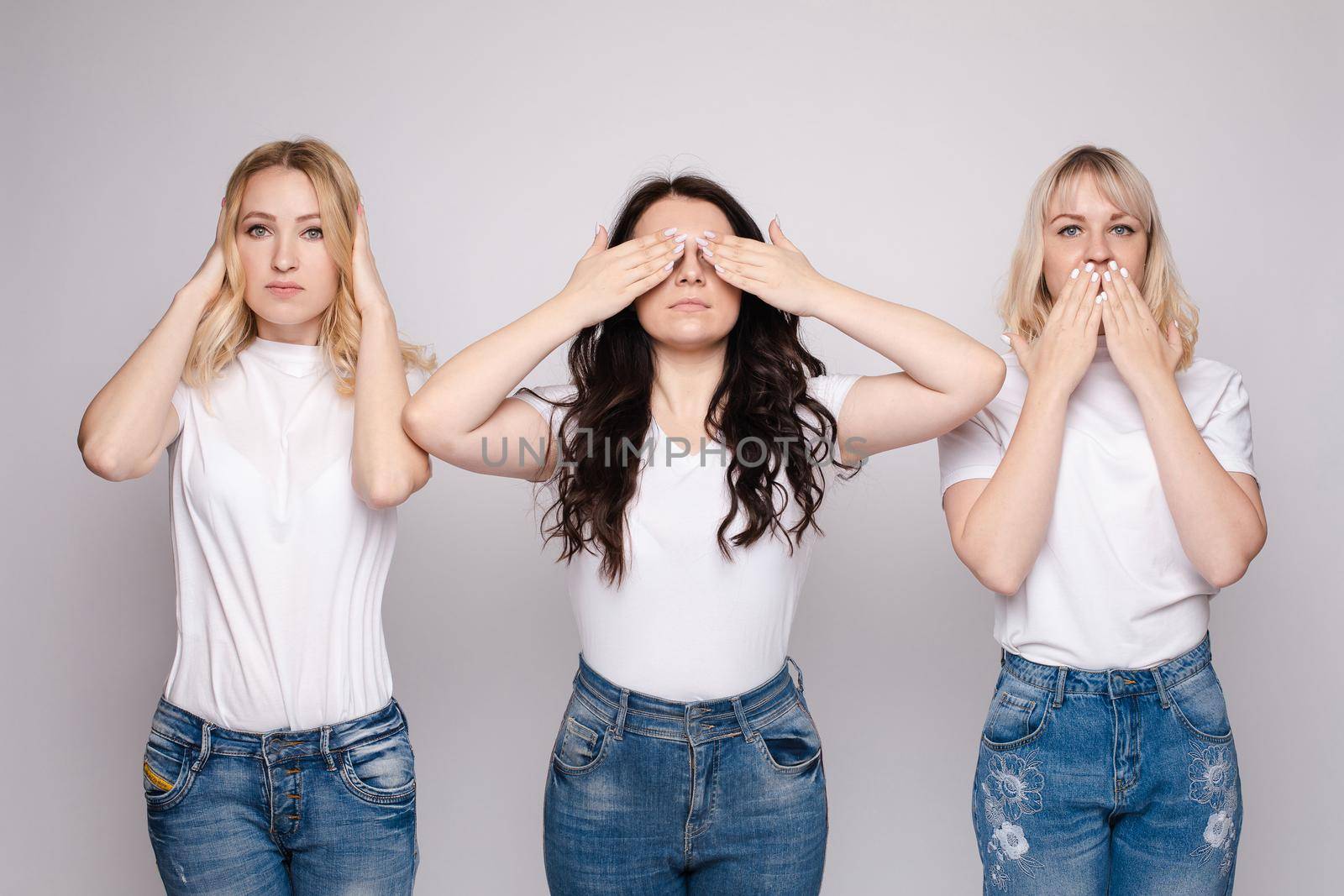 Crop of three young girls in casual clothes posing at camera. Women in white shirts and jeans hiding different parts of their faces. Brunette and blonde models showing famous concept of indifference.