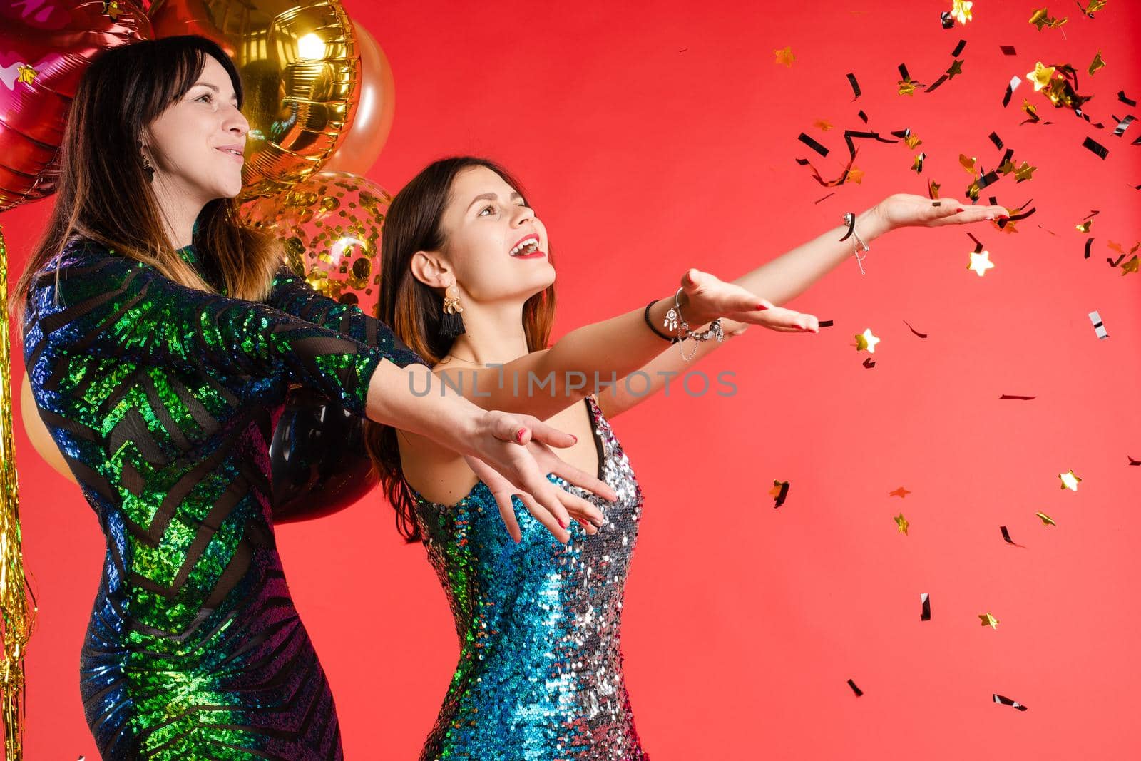 Group smiling beautiful woman posing surrounded by colorful shimmering confetti isolated red studio background. Happy three girl friend enjoying holiday party relaxing and having fun medium long shot