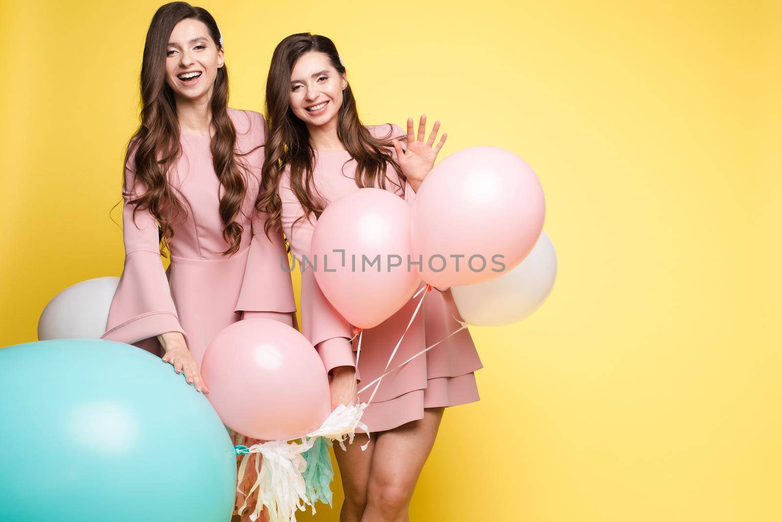 Studio portrait of gorgeous brunette twins posing in trendy elegant pink dresses. Stylish outlooks. Smiling at camera. Yellow background with mint and pink air balloons.