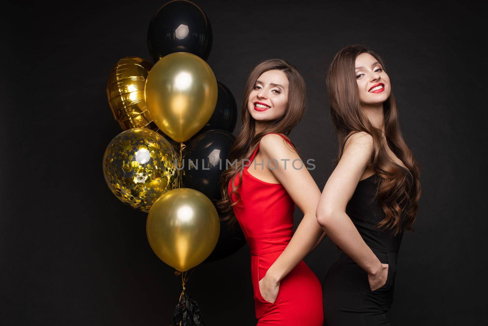 Cropped view of two beautiful sisters wearing red and black dresses and standing back to back in studio. Young long haired model posing near golden balloons, looking at camera and smiling on isolated.