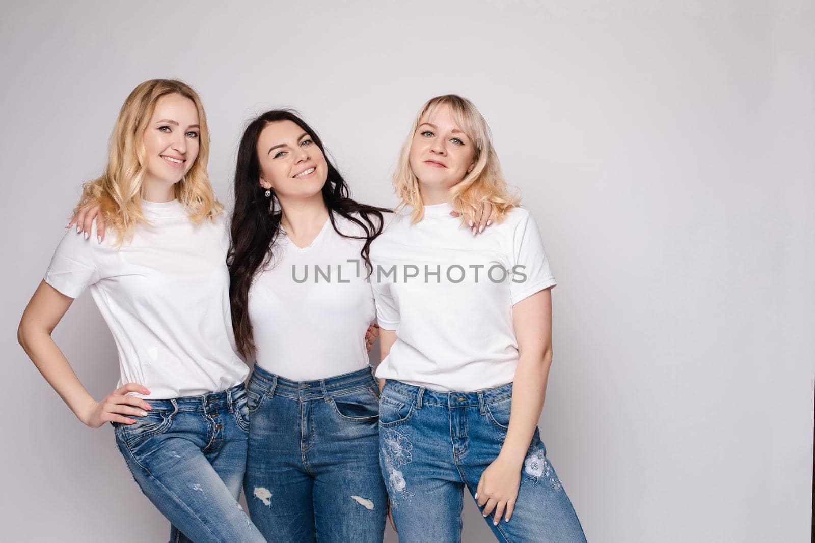 Front view of three beautiful women wearing white shirts and jeans and posing on isolated background. Stylish models standing in studio, looking at camera and smiling. Concept of casual street outfit.