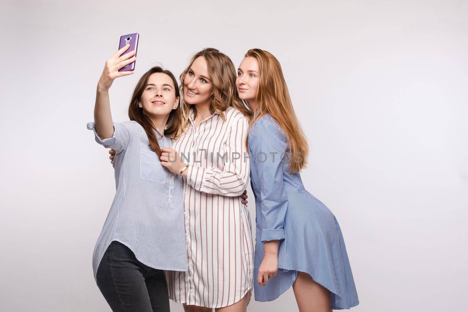 Studio portrait of stunning young girls in tops and denim taking self-portrait with smartphone. Isolate