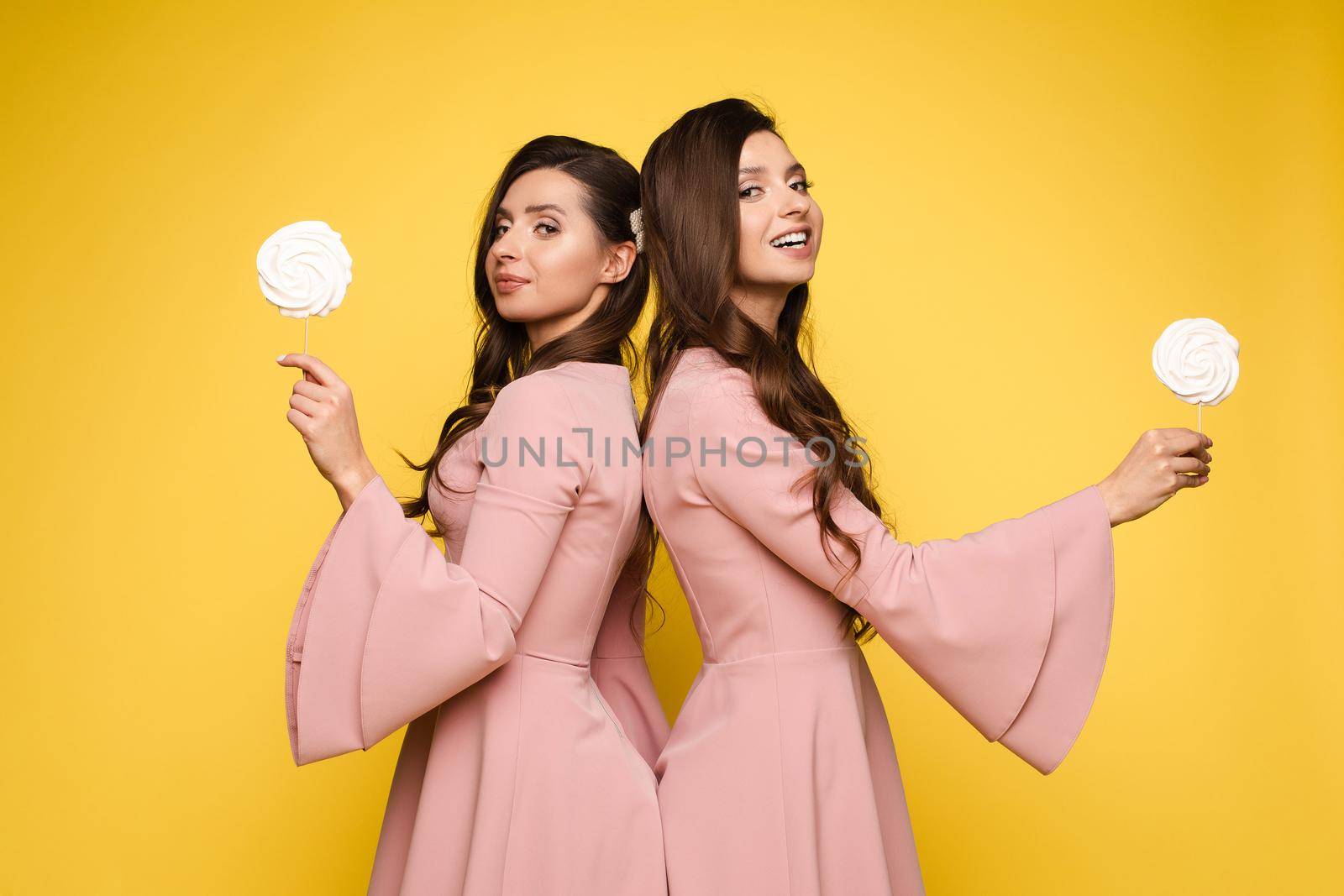 Front view of charming twins in pink dresses closing eyes with lollipops and posing on isolated background. Two female friends laughing and having fun in studio. Concept of childhood and beauty.